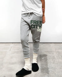 Someone wearing Free City FC Large 3/4 Sweats in Heather Surplus with white text, a white tee, cream slouchy socks, and black slippers stands on a concrete floor in front of a plain white wall.