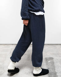 Someone wearing loose dark blue loungewear—a Free City Basic Goodness OG Sweatpant in Moonshrooms with a matching sweatshirt—plus white socks and black slip-ons walks indoors on a concrete floor.