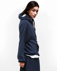A person with straight dark hair wears the Free City Basic Goodness Hoodie in Moonshrooms, paired with matching pants and a white shirt, standing against a plain white background and glancing over their shoulder.