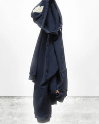 The Free City Basic Goodness Hoodie in Moonshrooms, an oversized navy blue style, hangs from its hood against a white wall. It features a white cloud on the hood and a colorful tag on the pocket for classic brand appeal.