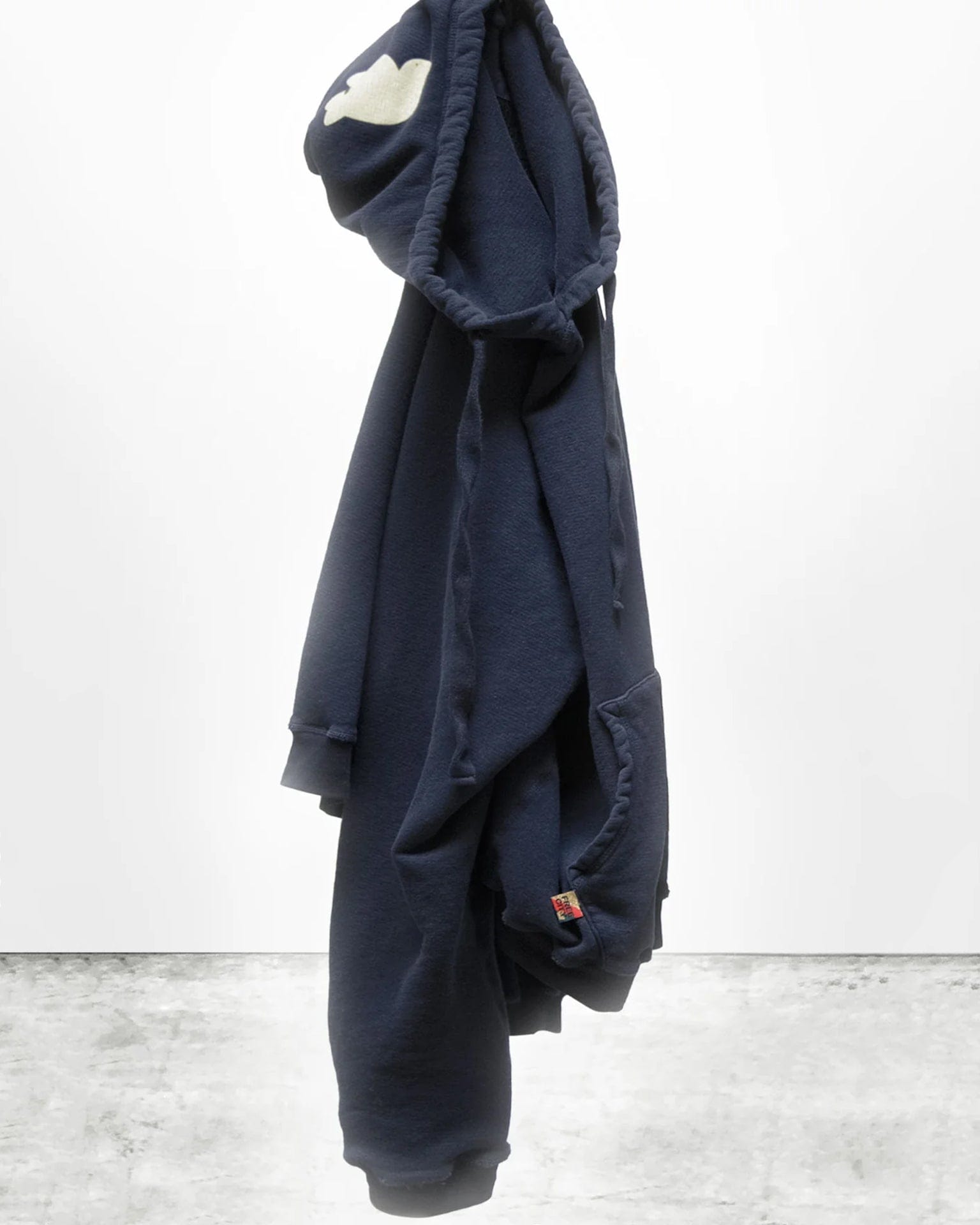 The Free City Basic Goodness Hoodie in Moonshroom Blue hangs against a white wall and concrete floor. Its hood, showing a white cloud patch, is up; the front pocket is visible. Made from soft light terry cotton for all-day comfort.