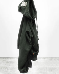 The Free City Basic Goodness Hoodie in Tent, a dark green oversized style with a white hood patch and twisted drawstrings, hangs against a plain white wall and concrete floor background, showcasing its relaxed fit.
