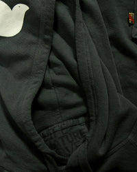Close-up of the Free City Basic Goodness Hoodie in Tent (dark green), showing a white bird graphic on the left, a pocket, a small orange tag, and partially visible printed text reading FUTURE.