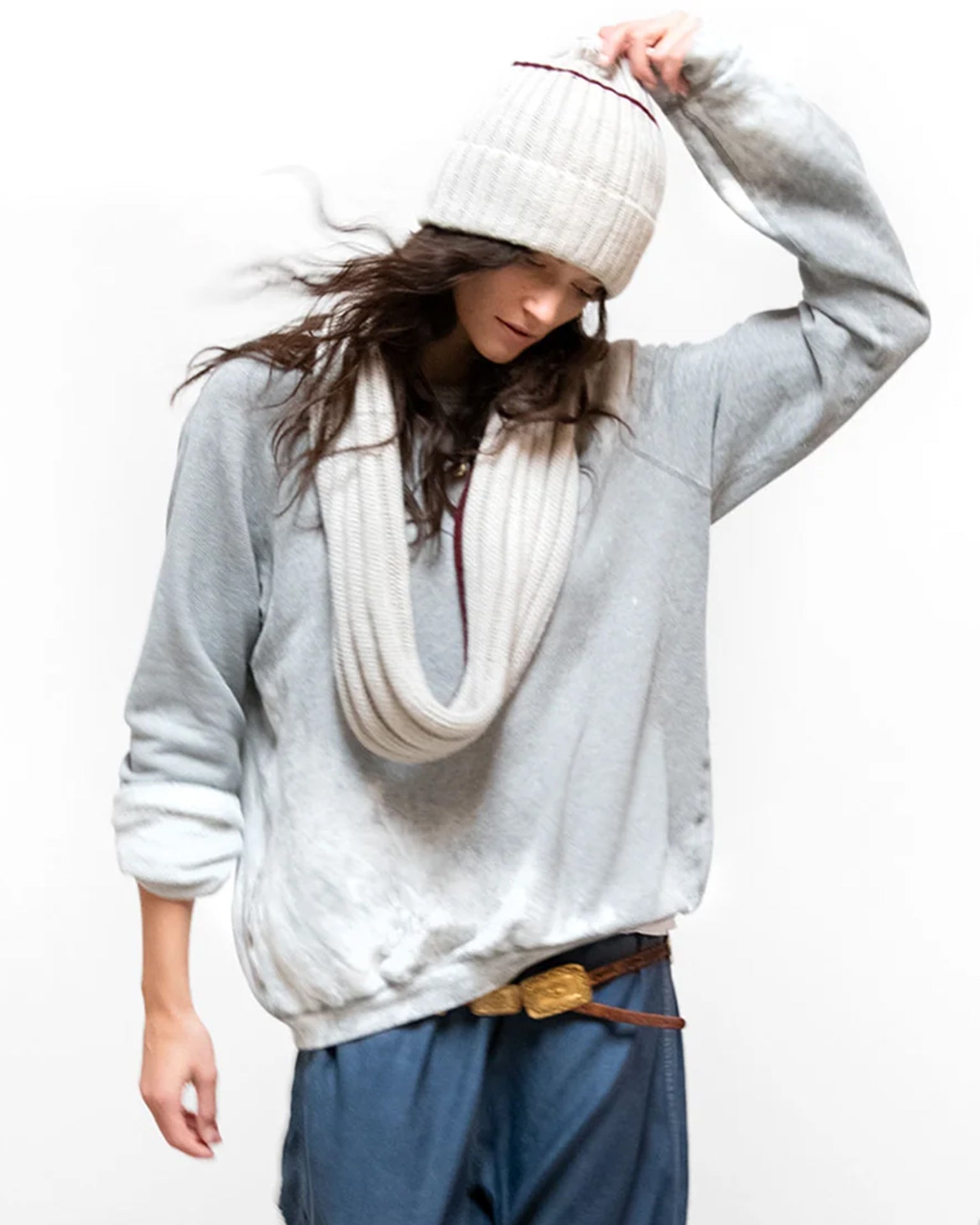 A person wearing a loose Free City Artistswanted Raglan in Heather Bleachout, blue pants, a cream knit beanie, and a matching scarf stands against a plain white background, looking down while adjusting the hat.