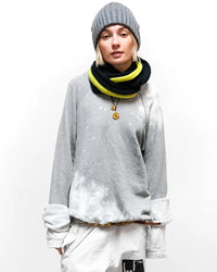 A person wearing a gray beanie, a Free City Rabbit Sweatshirt in Heather Bleachout, a gray and neon yellow scarf, white pants, and gold necklaces stands against a plain white background.