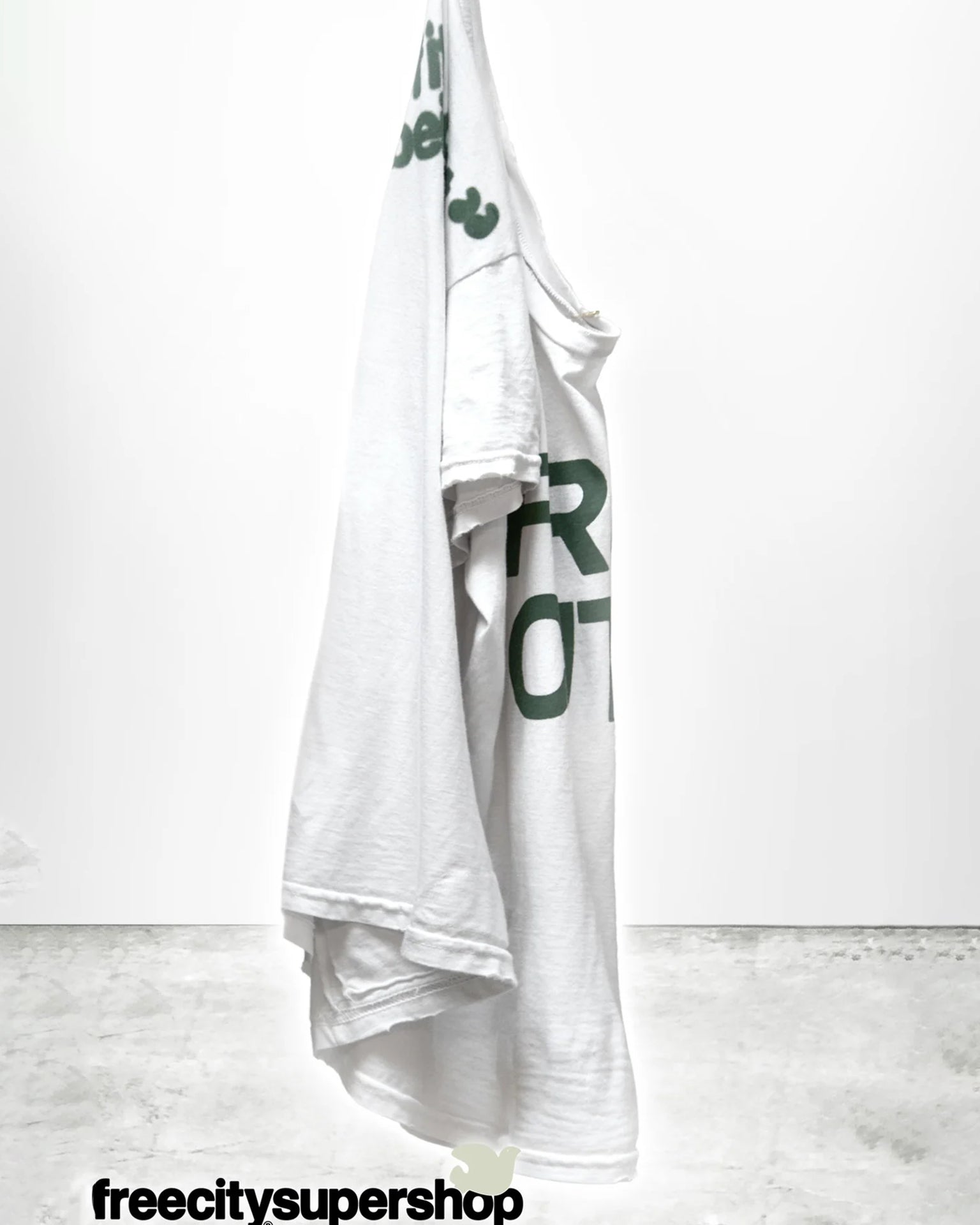 The Free City FC thedoingofitisthebeingofit Supervintage S/S Tee in Laundry Whites, featuring green text, hangs on a hook against a plain white background. "freecitysupershop" appears in black at the bottom of the image.