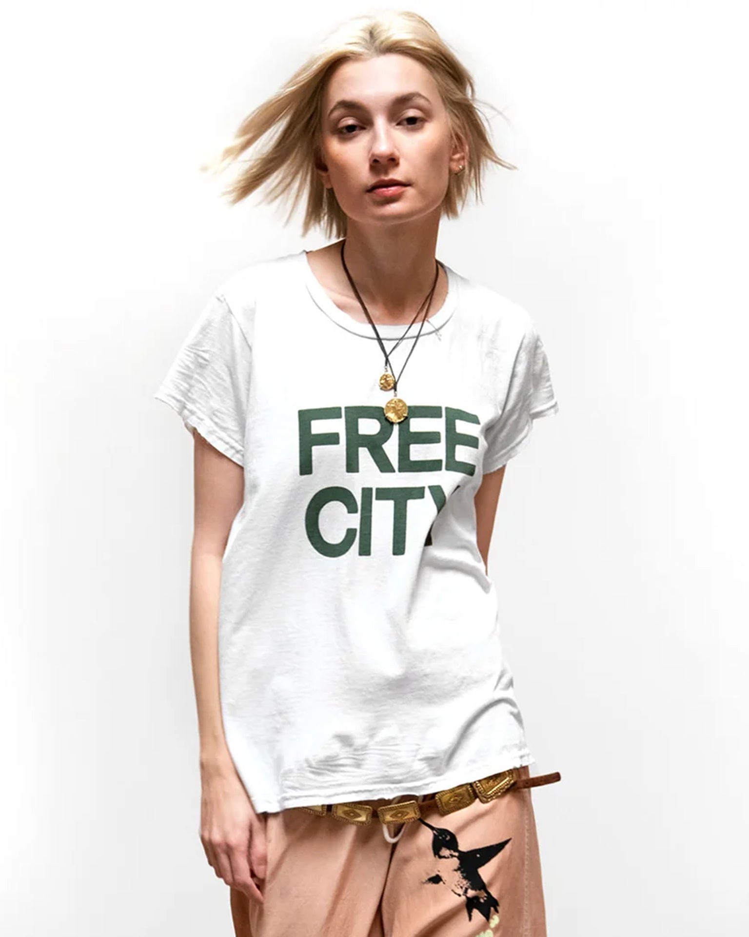 A person with short blonde hair wears a Free City FC thedoingofitisthebeingofit Supervintage S/S Tee in Laundry Whites, peach pants with a patterned belt, and a pendant necklace, standing against a plain white background.