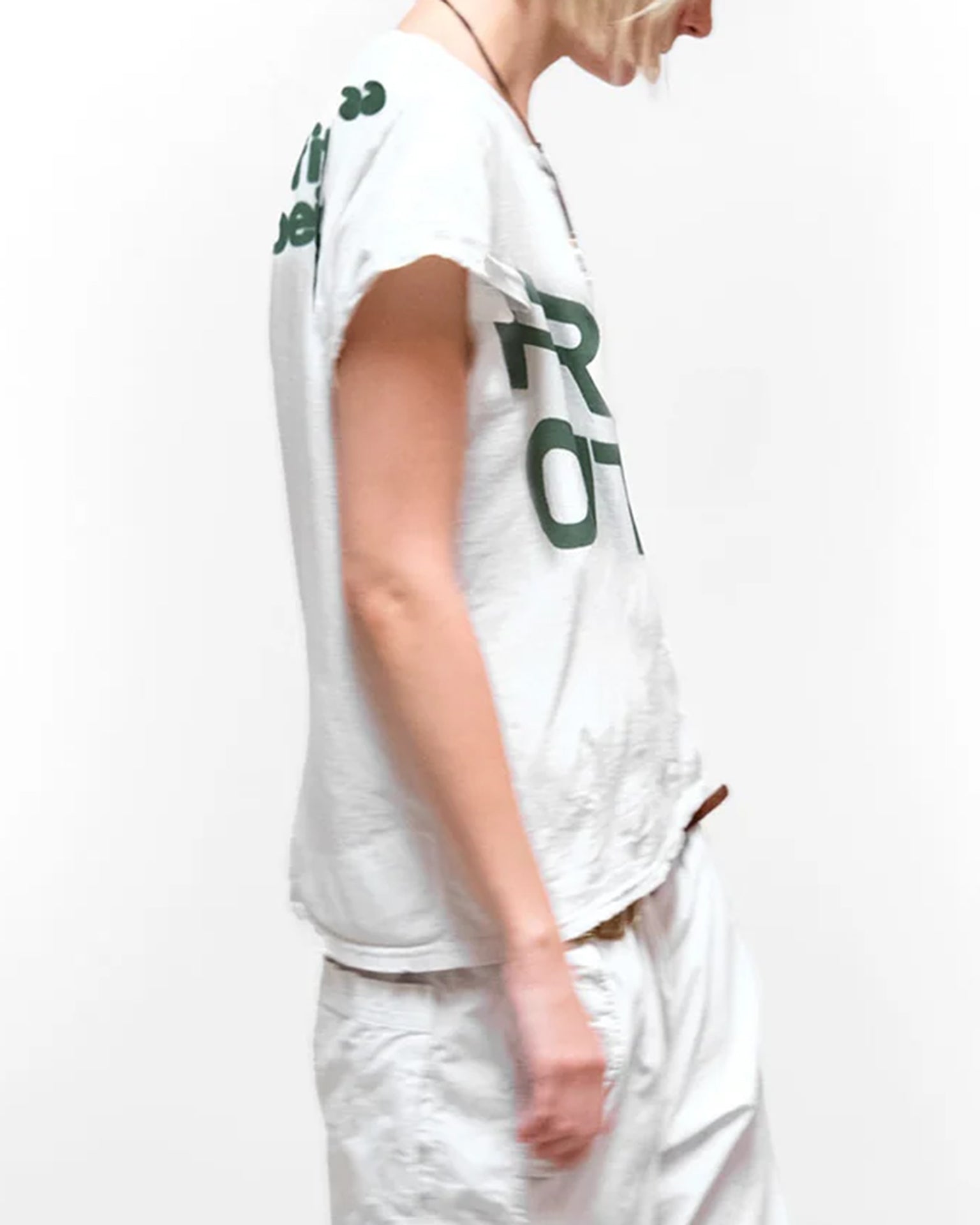 A person with short blonde hair wears the Free City FC thedoingofitisthebeingofit Supervintage S/S Tee in Laundry Whites and white pants, standing against a plain white background, facing slightly to the side with their arm down.