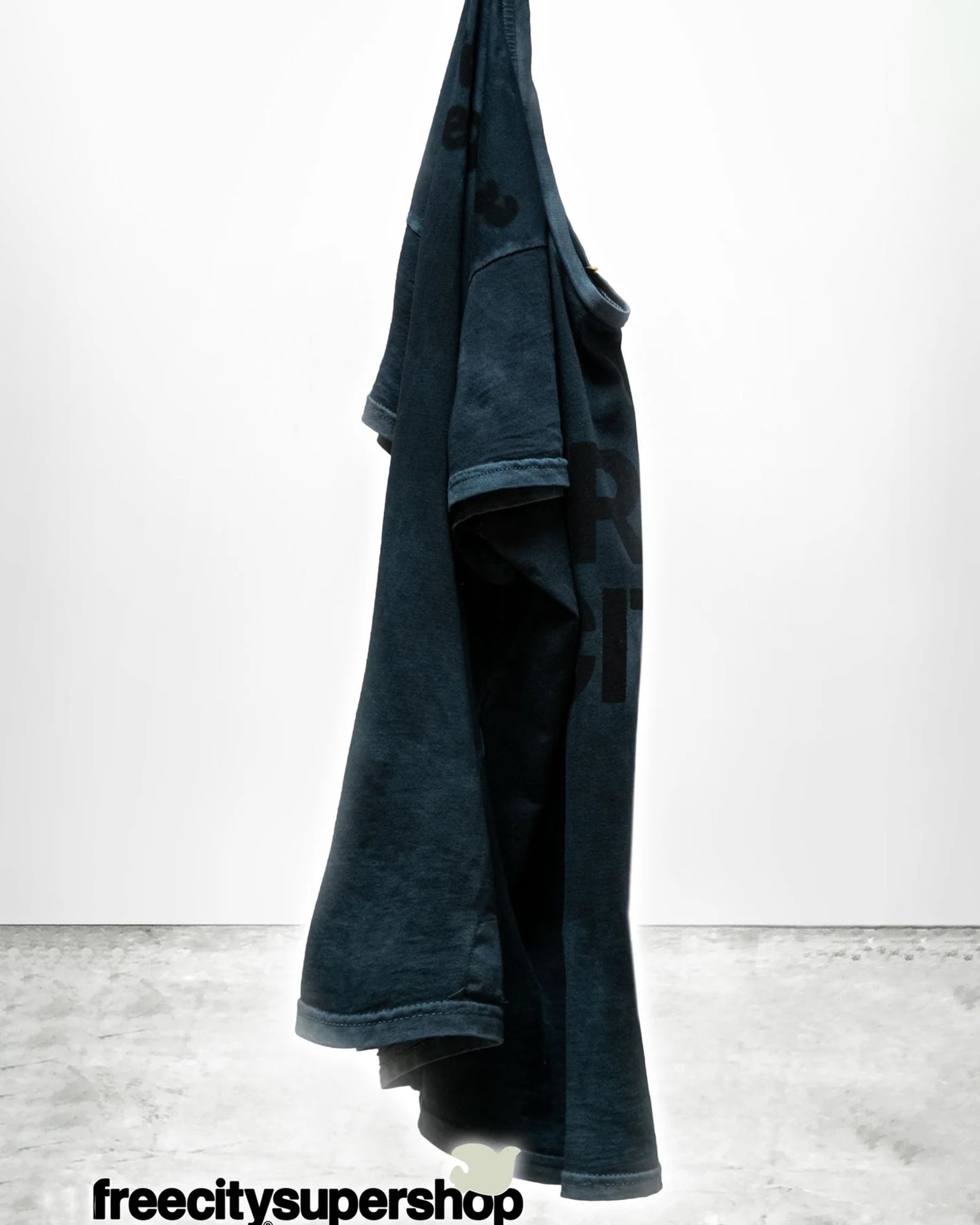 A dark blue FC thedoingofitisthebeingofit Supervintage S/S Tee in Volcano Powder by Free City hangs against a white background. “freecitysupershop” is in black text at the bottom on a gray concrete floor.