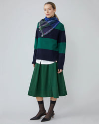 A woman stands against a plain background wearing a navy and green striped sweater, green skirt, white shirt, Moismonts Chai Wool Square Scarf No 752 in Royal Blue, sheer black socks, and black heels.