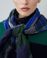 A woman wears a Moismont Chai Wool Square Scarf No 752 in Royal Blue, tied at the neck over a green and navy sweater. Only the lower half of her face is visible.