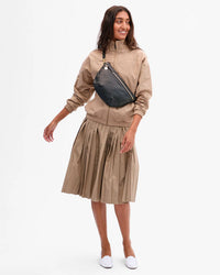 A woman in a beige zip-up jacket and pleated skirt smiles, wearing the Clare V. Grande Fanny - Petit Caviar in Black crossbody and white slip-on shoes against a plain white background.