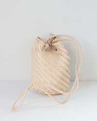 The Pom Pom Double Jump in Peachy Natural by Dragon Diffusion is a small, light-colored crossbody straw bag with a drawstring closure and two braided shoulder straps, shown on a plain white surface against a white background.