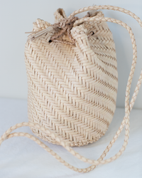 A close-up of the Dragon Diffusion Pom Pom Double Jump in Peachy Natural, a mini woven leather bucket bag with a drawstring closure and long braided crossbody straps, displayed on a white surface.