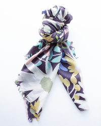 The Bold Daisy Wool Silk & Cashmere Scarf in Plum by Épice features a hand-printed floral design and frayed edges, styled in a loose knot against a white background.