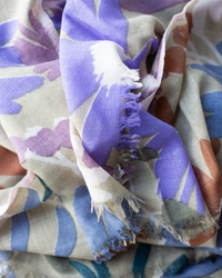 A close-up of Épice’s Bold Daisy Wool Silk & Cashmere Scarf in Taupe shows its frayed edge and floral design in purple, blue, and beige tones.