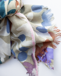 A close-up of Épices Bold Daisy Wool Silk & Cashmere Scarf in Taupe, featuring a lightweight design with fringe and an abstract floral pattern in blue, green, beige, brown, and purple on white.