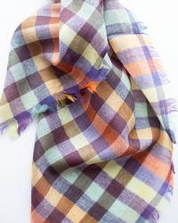 The Épice Plaid Wool Bandana in Orange features a multicolored checkered pattern with purple, orange, brown, and light green shades and a frayed edge.