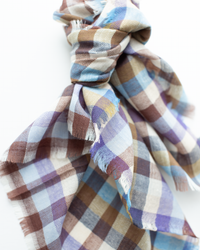 A close-up of the Épice Plaid Wool Bandana in Blue, featuring frayed edges and blue, brown, and beige tones, tied loosely against a white background.