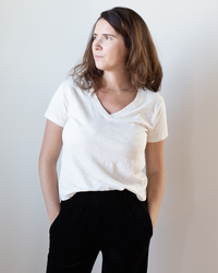 A woman with long brown hair, wearing the Velvet by Graham & Spencer Eli V Neck Top in Bisque and black pants, stands with her hands in her pockets against a plain light background, looking to the side.