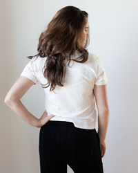 A person with long brown hair, seen from behind, wears the Velvet by Graham & Spencer Eli V Neck Top in Bisque and black pants, one arm on their hip against a plain light background.