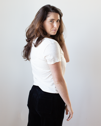 A woman with long brown hair wears the Eli V Neck Top in Bisque by Velvet by Graham & Spencer and black pants, standing slightly turned to the side and looking over her shoulder toward the camera, set against a plain light background.