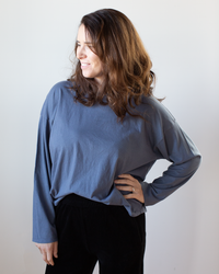 A woman with wavy brown hair, wearing the NSF Loffler Boxy Fine Jersey Tee in Indigo and black pants, stands smiling with one hand on her hip against a plain light background.