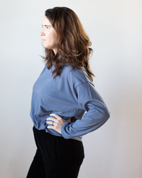 A woman with long brown hair stands in profile against a light background, wearing the NSF Loffler Boxy Fine Jersey Tee in Indigo and black pants, one hand on her hip and a neutral expression.