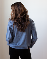 A woman with long, wavy brown hair faces away from the camera, wearing an NSF Loffler Boxy Fine Jersey Tee in Indigo and black pants against a plain light background.