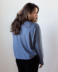 A woman with long brown hair, wearing the NSF Loffler Boxy Fine Jersey Tee in Indigo and black pants, stands slightly turned away from the camera against a plain light background.