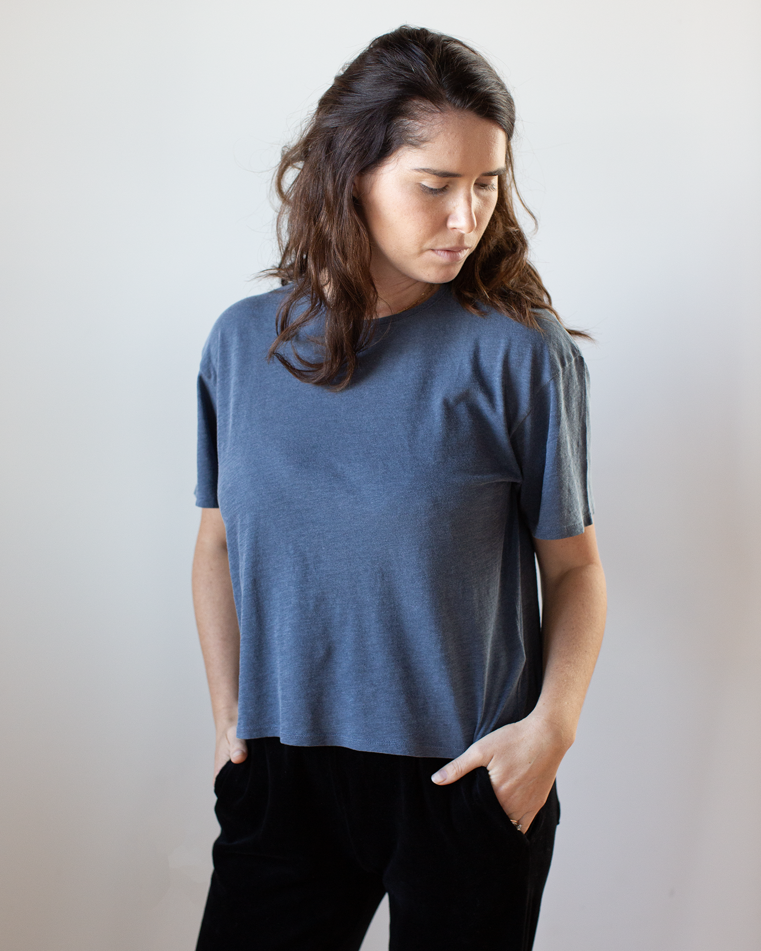 A woman with long brown hair stands against a plain backdrop, wearing the NSF Harper Boxy Slub Jersey Tee in Faded Indigo and black pants, hands in pockets as she gazes down and to the side.