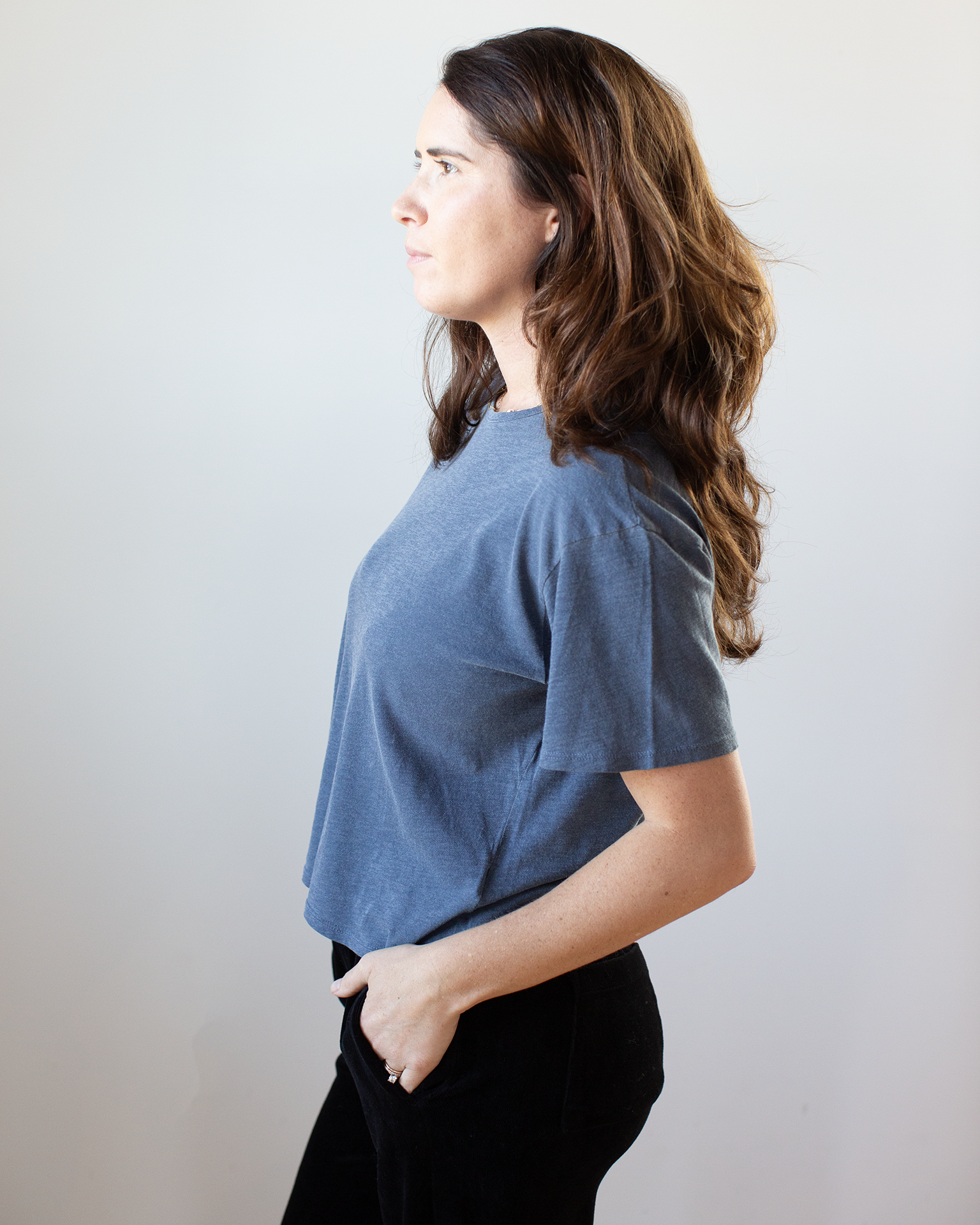 A woman with long brown hair stands in profile against a light background, wearing the NSF Harper Boxy Slub Jersey Tee in Faded Indigo and black pants, one hand in her pocket.