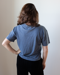 A person with medium-length brown hair faces away, wearing the NSF Harper Boxy Slub Jersey Tee in Faded Indigo and black pants, one hand on their hip, set against a neutral background.