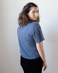 A woman with wavy brown hair models the NSF Harper Boxy Slub Jersey Tee in Faded Indigo, paired with black pants, standing sideways and looking at the camera against a plain light background.