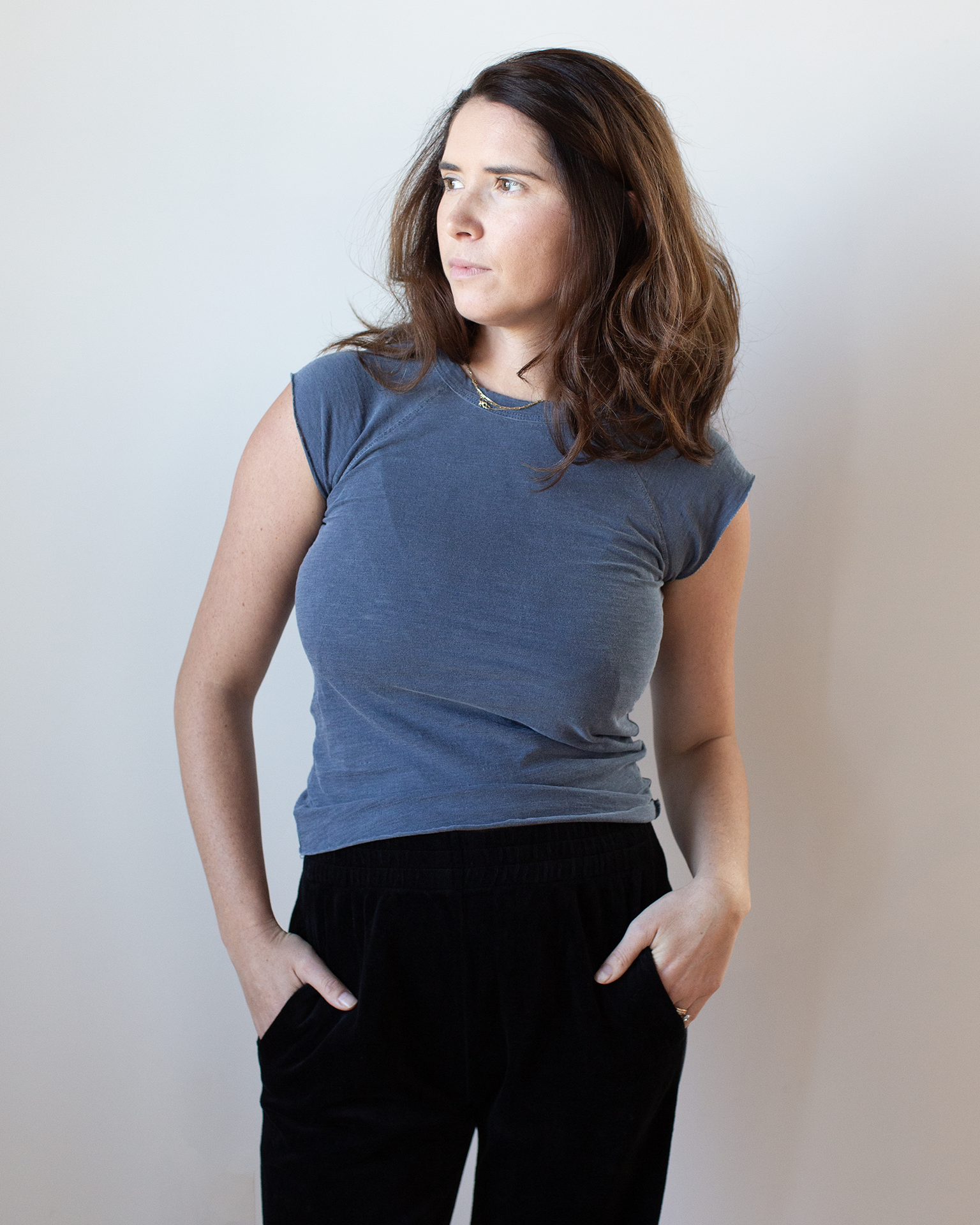 A woman with long brown hair wears the NSF Chloe Fitted Slub Jersey Muscle Tee in Faded Indigo and black pants, standing against a light background with her hands in her pockets, looking to the side with a neutral expression.