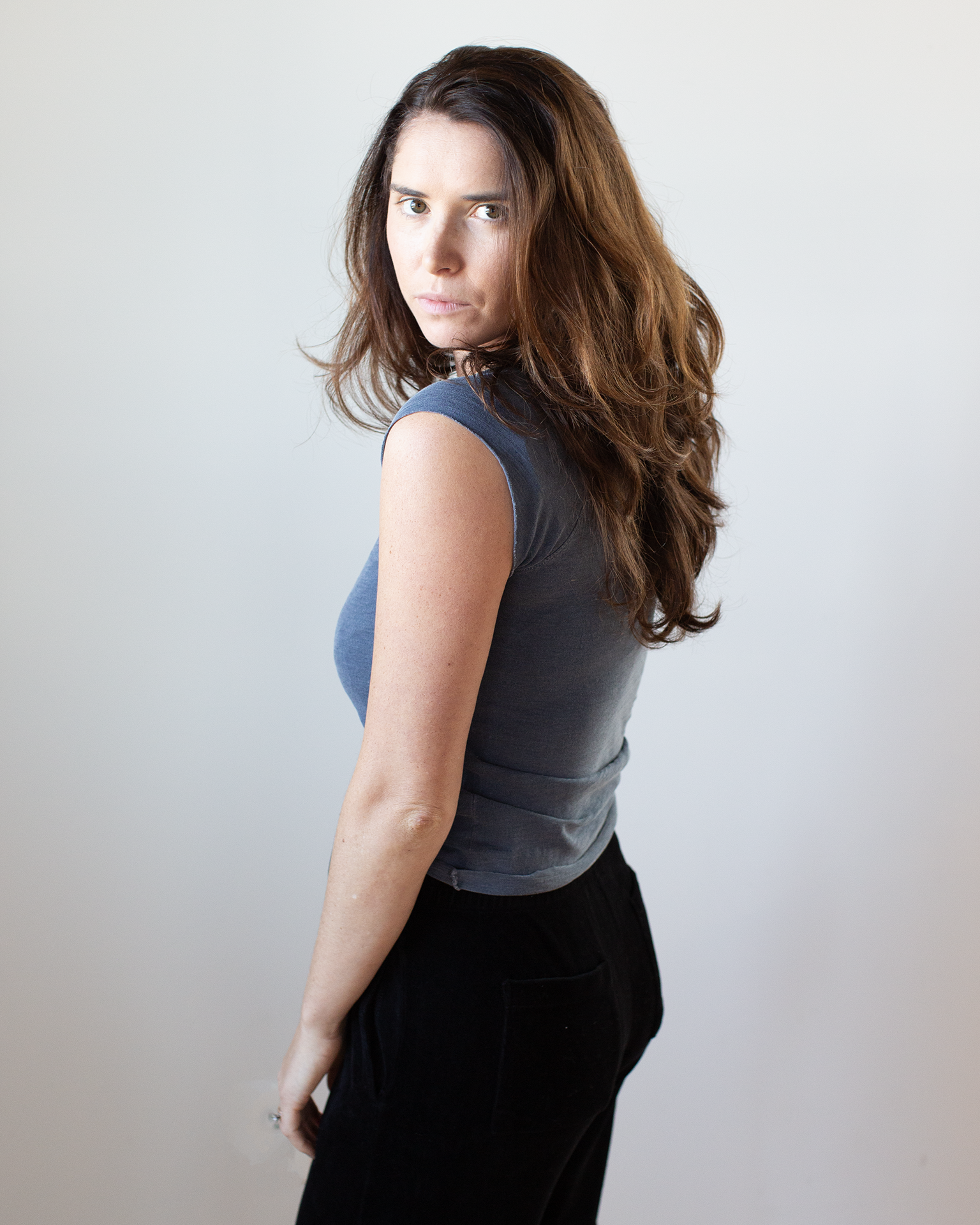 A woman with long brown hair stands sideways against a plain background, wearing the NSF Chloe Fitted Slub Jersey Muscle Tee in Faded Indigo and black pants, gazing seriously at the camera.