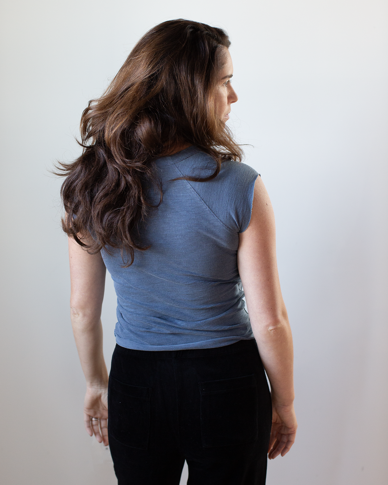 A woman with long brown hair wears the NSF Chloe Fitted Slub Jersey Muscle Tee in Faded Indigo and black pants, standing with her back to the camera against a plain light background.