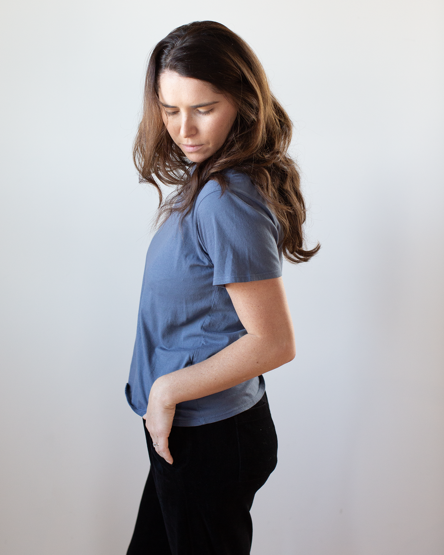 A woman with long brown hair stands sideways, hand in pocket and head tilted down, wearing the NSF Moore Relaxed Fine Jersey Tee in Indigo and black pants against a plain light background.