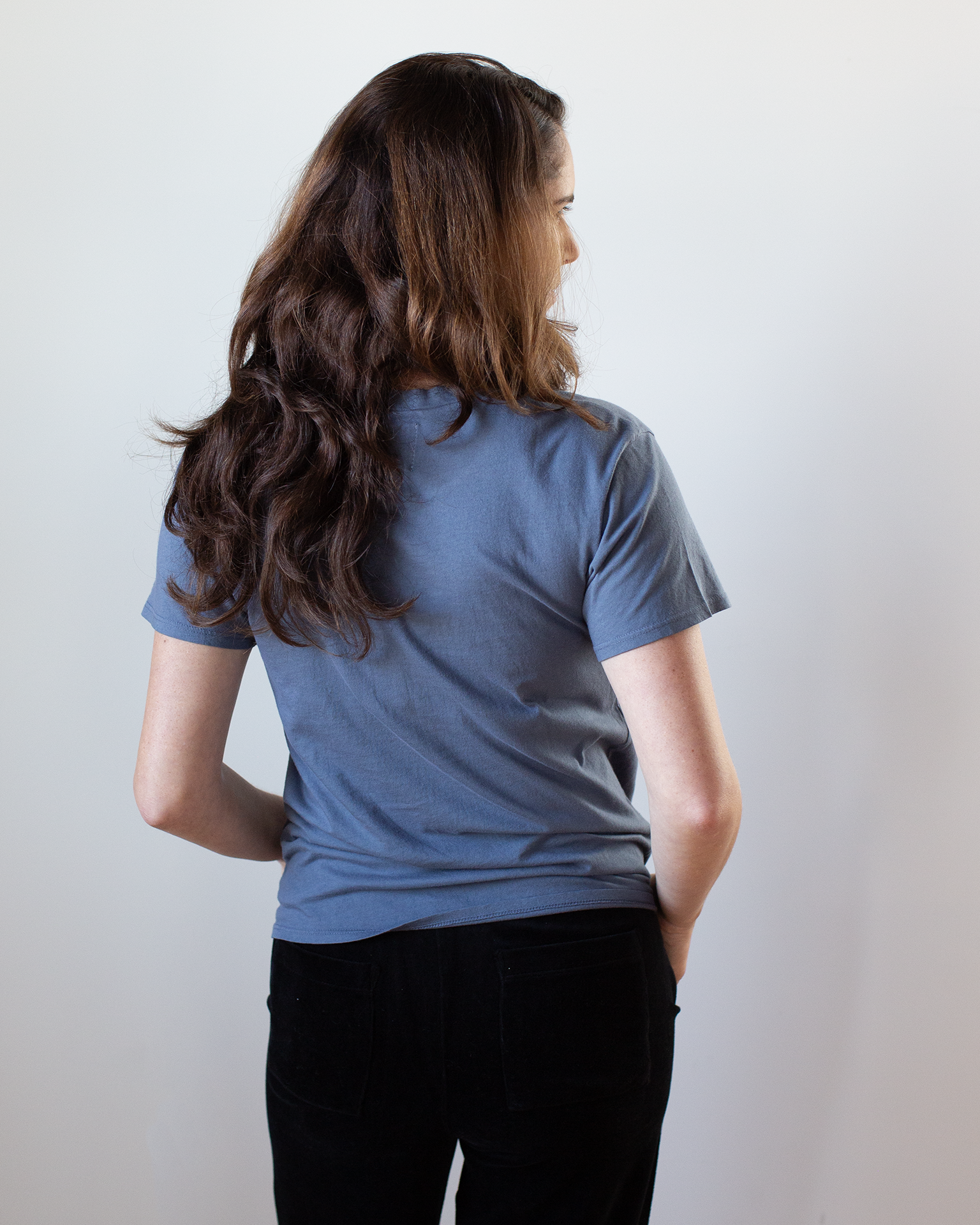 A person with long brown hair faces away from the camera, wearing the NSF Moore Relaxed Fine Jersey Tee in Indigo and black pants against a plain white background.