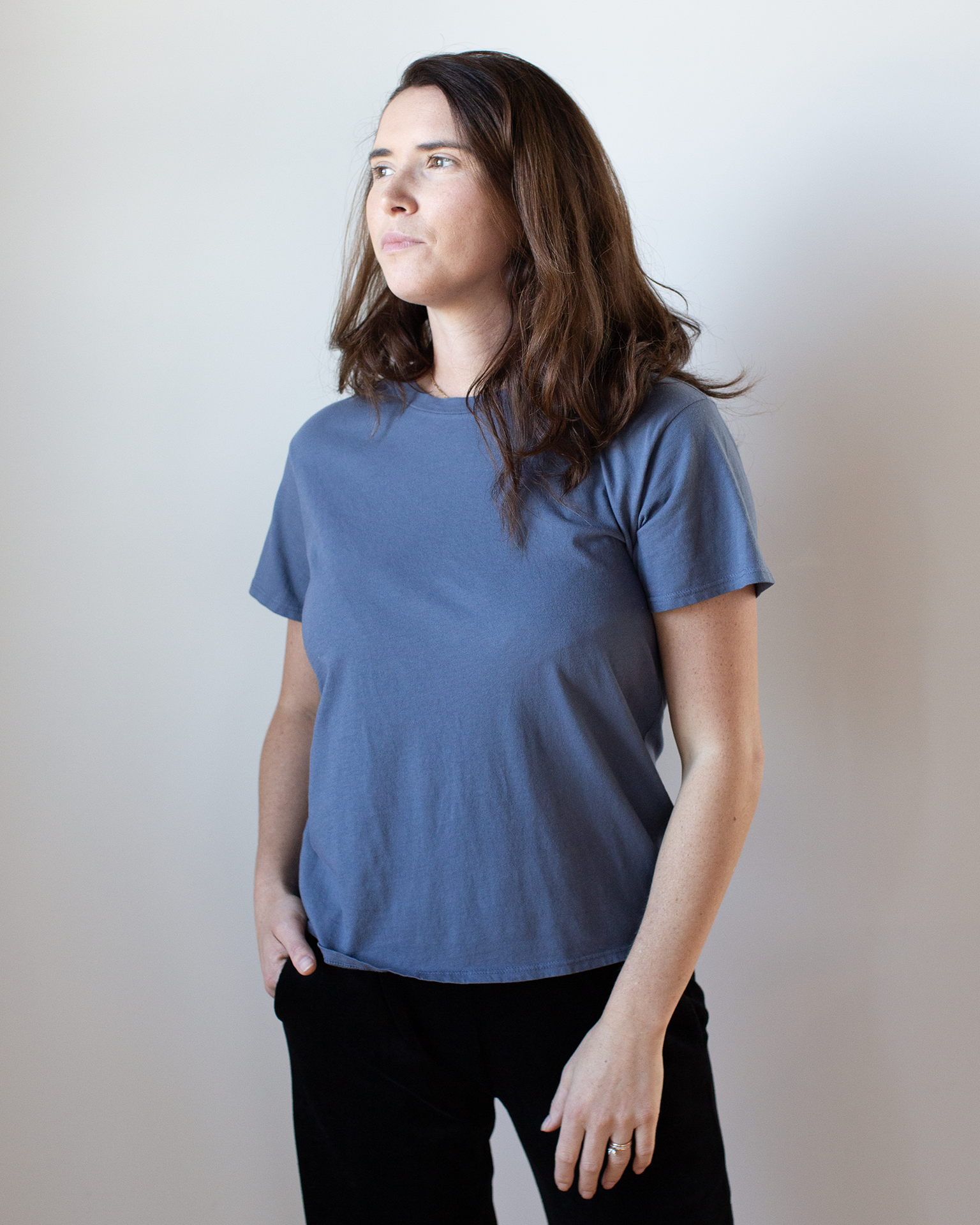 A person with long brown hair stands against a light background, looking slightly to the side with one hand in their pocket, wearing the NSF Moore Relaxed Fine Jersey Tee in Indigo and black pants.