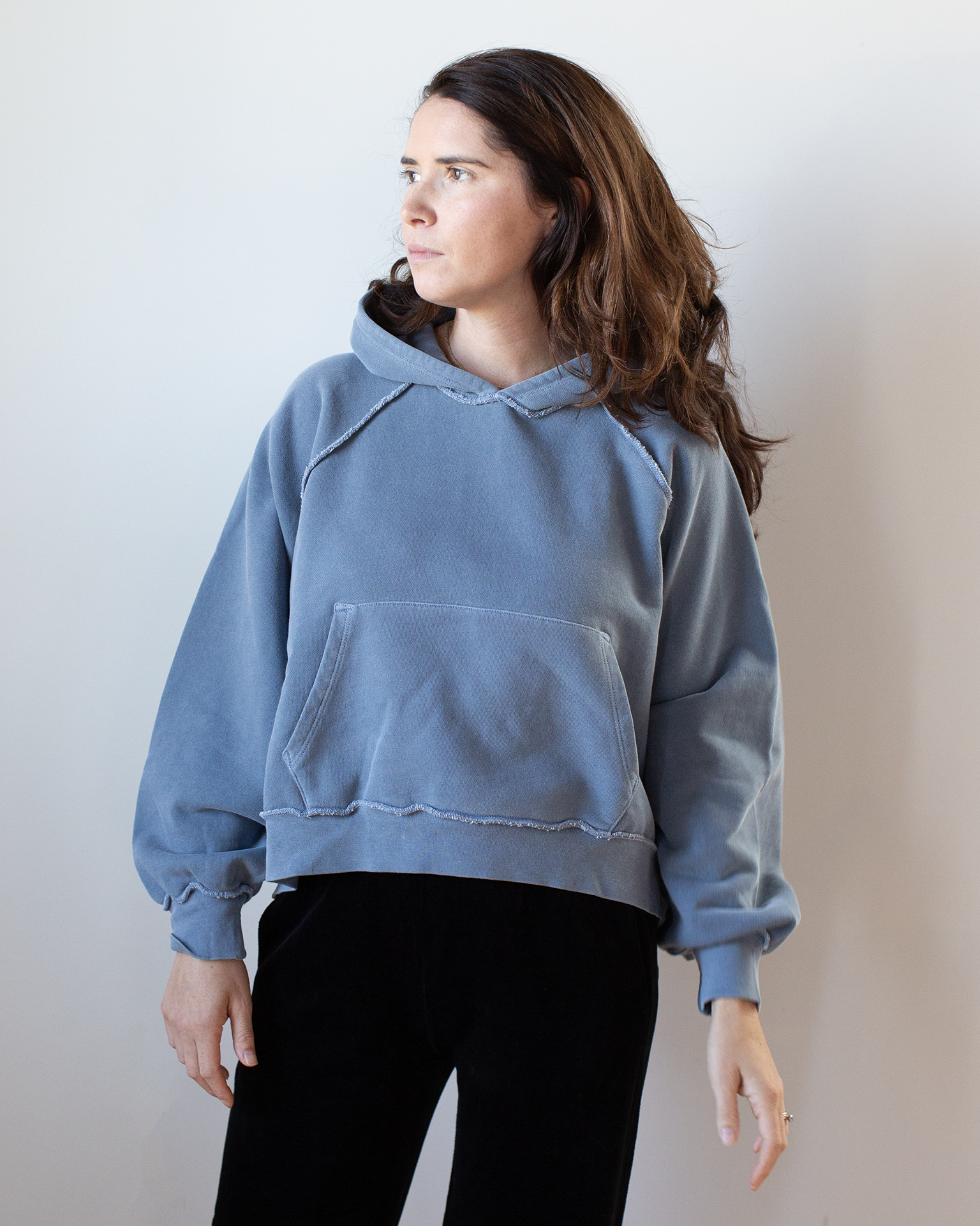 A person with long brown hair wears the NSF Luma Oversize FT Poncho in Faded Indigo and black pants, standing against a light background and looking left with a neutral expression.