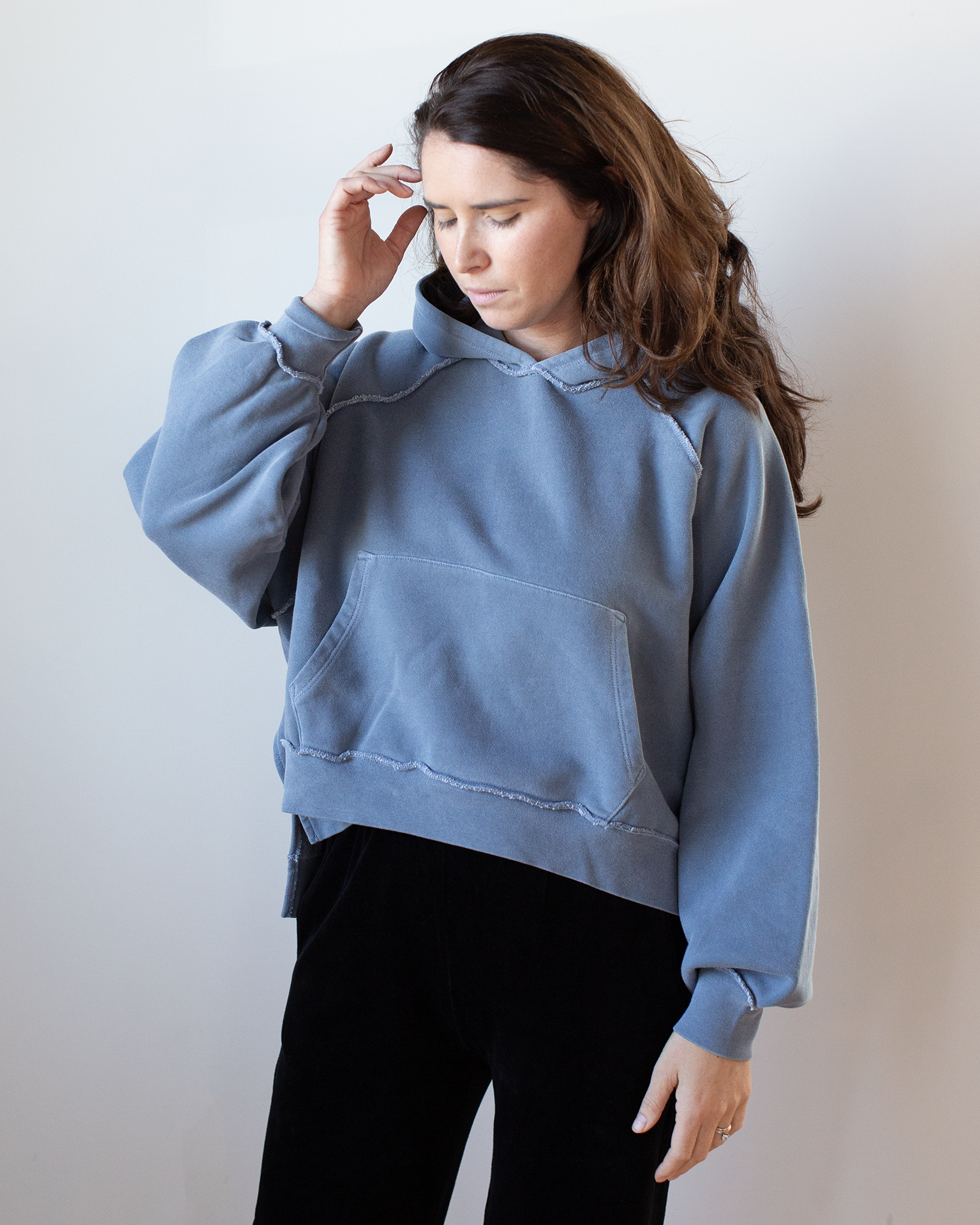 A person with long brown hair wears the NSF Luma Oversize FT Poncho in Faded Indigo and black pants, looking down and touching their hair against a plain light background.