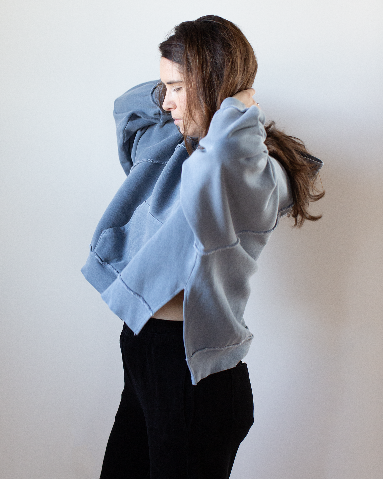 A woman with long brown hair, wearing the NSF Luma Oversize FT Poncho in Faded Indigo and black pants, stands sideways against a plain white background, adjusting her hair with both hands.