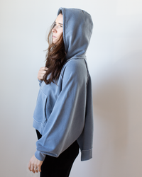 A person stands sideways against a plain light background, wearing the NSF Luma Oversize FT Poncho in Faded Indigo, gazing ahead with one hand holding the hoodie near their chest.
