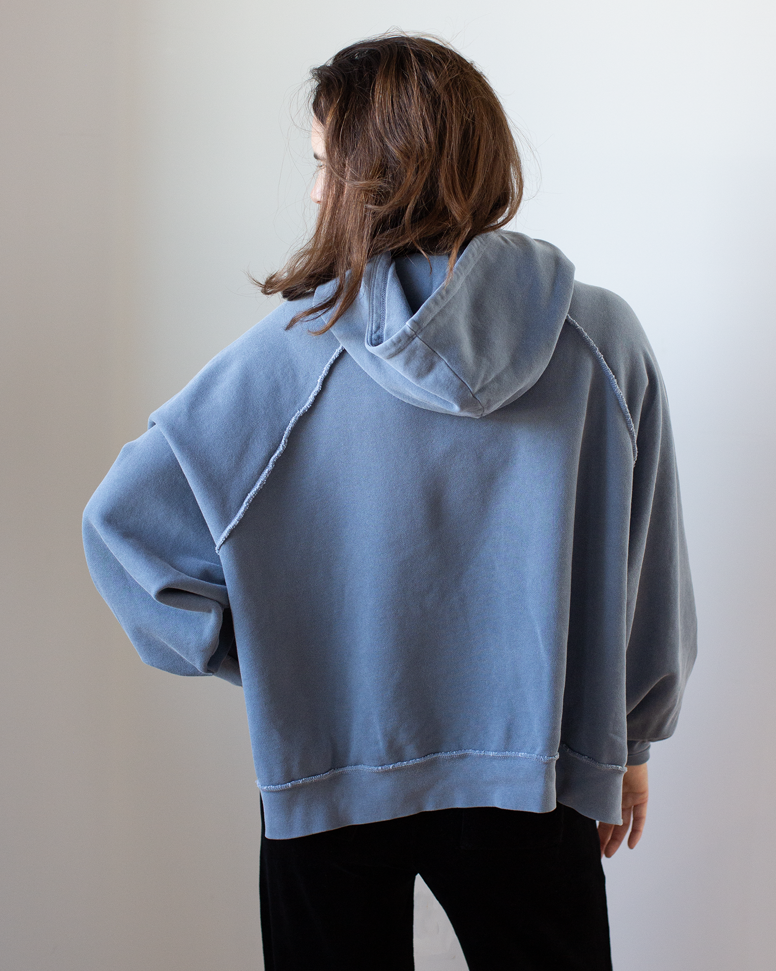 A person with long brown hair faces away, wearing the NSF Luma Oversize FT Poncho in Faded Indigo and black pants against a plain light background.