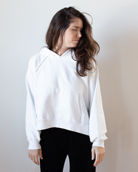 A woman with long brown hair, wearing an oversized NSF Luma Oversize FT Poncho in Chalk and black pants, stands against a plain light background, looking down and slightly to her right.
