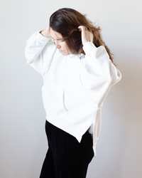 A person with long brown hair wears the NSF Luma Oversize FT Poncho in Chalk and black pants, standing against a plain light background while looking down and adjusting their hair with both hands.