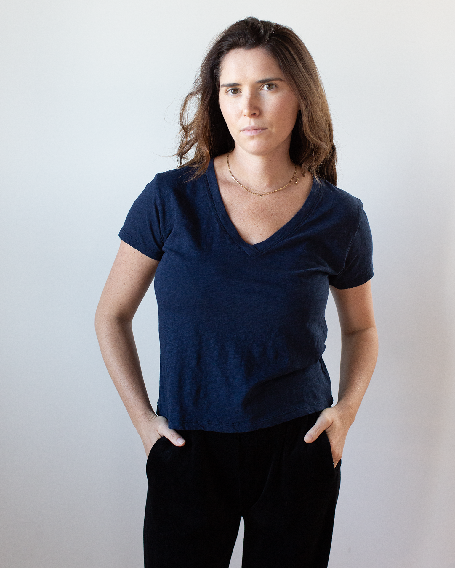 A woman with long brown hair wears the Eli V Neck Top in Midnight by Velvet by Graham & Spencer and black pants, standing against a plain light background with her hands in her pockets, looking at the camera.