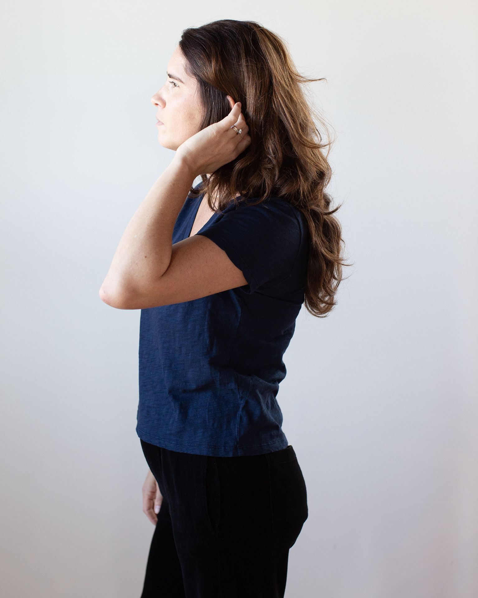 A woman with long brown hair stands in profile, touching her ear, wearing the Eli V Neck Top in Midnight by Velvet by Graham & Spencer and black pants against a plain light background.