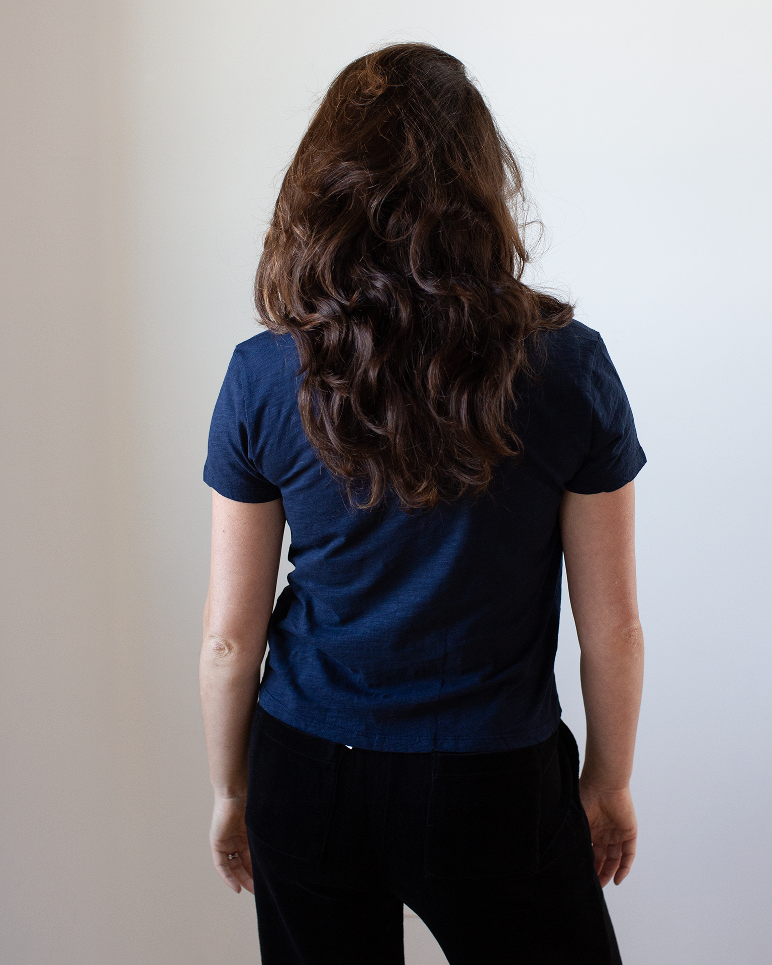 A woman with wavy brown hair stands facing away from the camera, wearing the Velvet by Graham & Spencer Eli V Neck Top in Midnight and black pants, her arms relaxed at her sides against a plain light background.